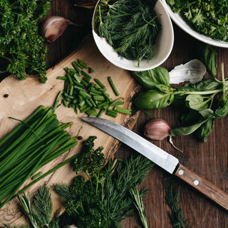 A wooden cutting board topped with an assortment of fresh herbs, including chives, parsley, dill, and basil. A knife rests on the board, alongside some chopped chives. Garlic cloves and additional herbs are scattered around the board, creating a rustic and vibrant culinary scene. The arrangement emphasizes the freshness and variety of the herbs.