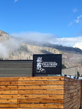 The exterior of Telluride Garden Center with a wooden fence in the foreground and a sign featuring the logo and name of the garden center. The backdrop includes a view of misty mountains under a blue sky with scattered clouds, highlighting the scenic location of the garden center.