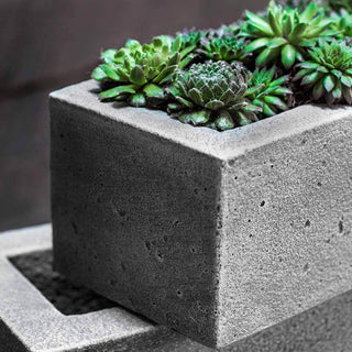 A close-up of a square concrete planter filled with green succulents. The planter has a minimalist design with sharp edges and a textured grey surface, contrasting with the vibrant green of the plants.