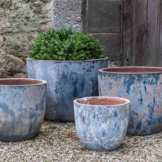 A collection of large, weathered blue-gray planters set against a stone wall. One of the planters contains a lush green shrub, while the others are empty. The scene captures a rustic and natural aesthetic, highlighting the textured surfaces of the planters and the greenery within the filled one. The planters are placed on a gravel surface, adding to the overall earthy and serene ambiance.