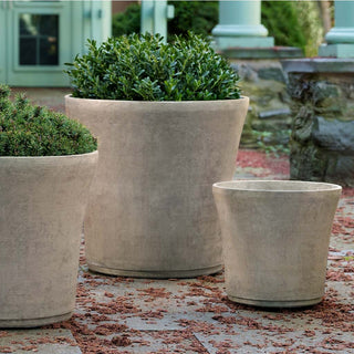 Three large, grey planters of different sizes are arranged on a patio. The planters contain lush green plants, adding a touch of greenery to the outdoor space. The background features stone walls and pillars, suggesting a garden or courtyard setting.