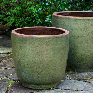 Two large green ceramic planters with a rustic finish, placed on a stone-paved area with greenery in the background.