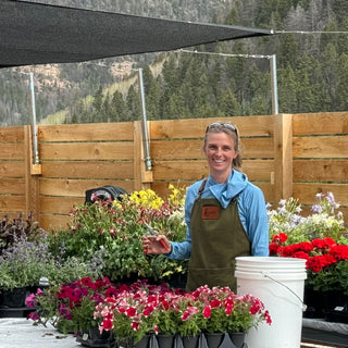 llison Ramsay, wearing an apron and holding pruning shears, stands smiling among a variety of colorful flowers and plants. The scene is set outdoors with wooden fencing and a backdrop of green trees and mountains, highlighting a vibrant and cheerful gardening environment.