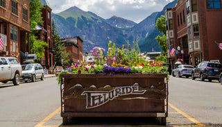 A vibrant street scene in Telluride, with colorful flowers planted in a large wooden planter bearing the town's name. The planter is placed in the middle of the street, with historic brick buildings, American flags, and parked cars lining both sides. Majestic mountains and a partly cloudy sky form a stunning backdrop, capturing the charm and beauty of this mountain town.