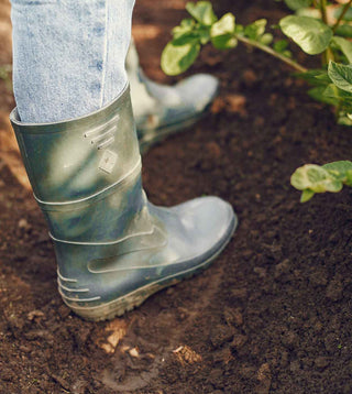 A close-up of a person wearing green rubber boots and jeans, standing on rich, dark soil in a garden, with green plants visible in the background.