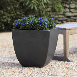 A black, rectangular planter with blooming blue flowers, placed next to a wooden bench on a gravel surface.